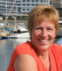 Eine Frau mit kurzen blonden Haaren und einem roten T-Shirt sitzt lächelnd an einem Hafen